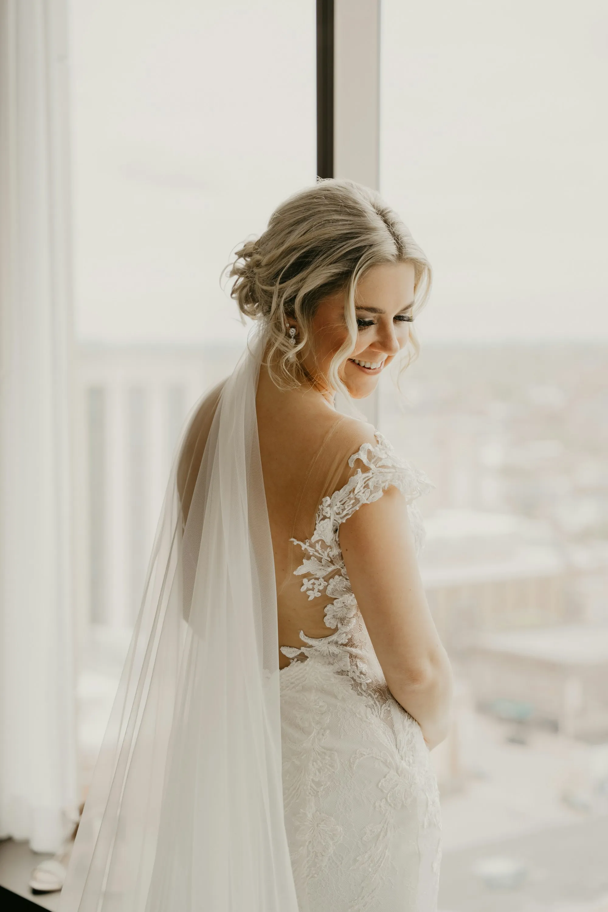 Bride in a lace gown and veil smiling by a window